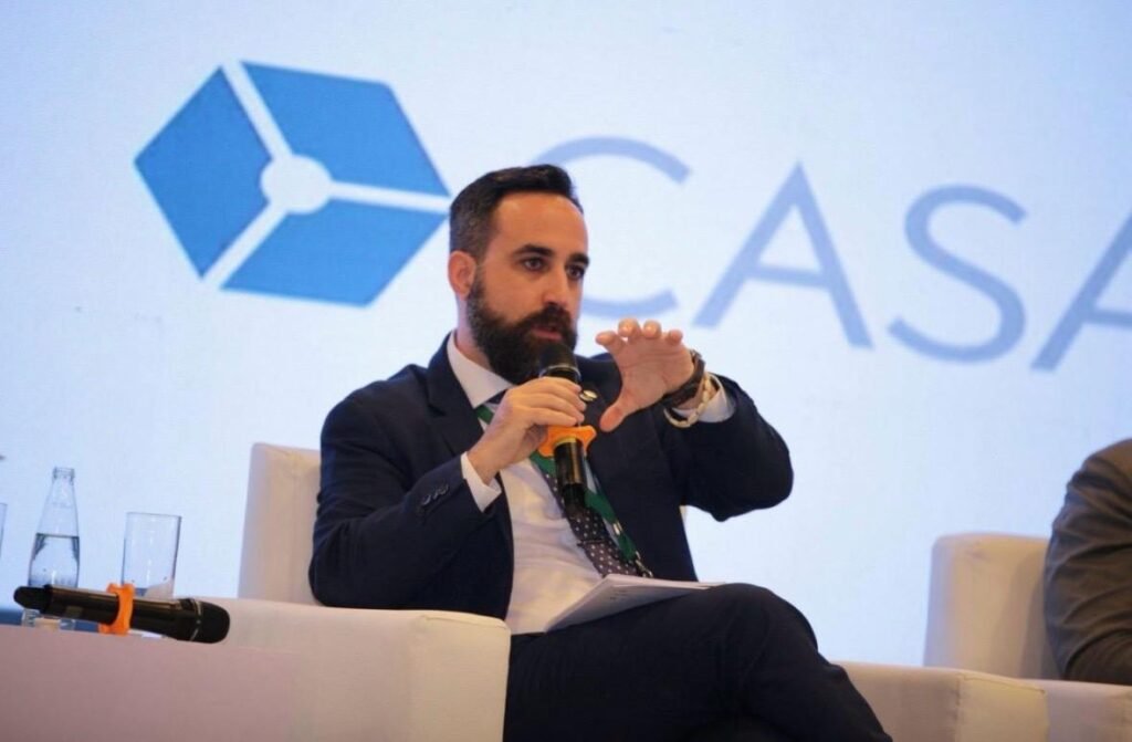 Rafael Mendías, con traje azul marino, interviniendo en primer plano con el micrófono en la mano derecha y la izquierda abierta en gesto de explicar, ante un fondo blanco con el logotipo de CASA