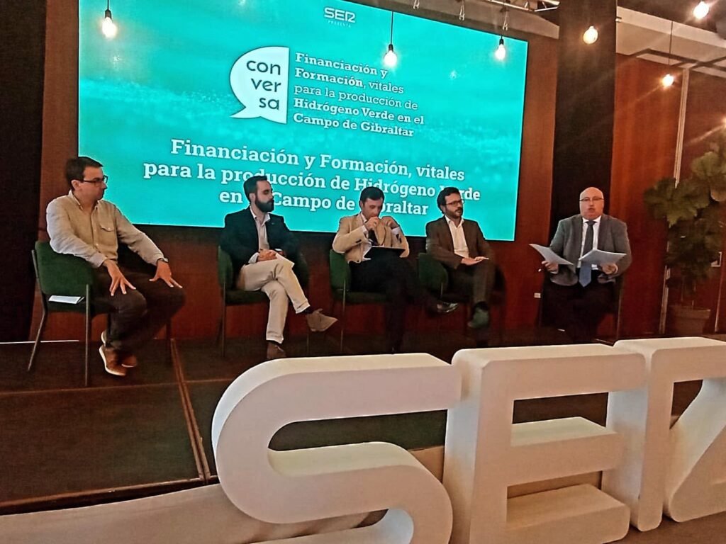 Mesa redonda de cinco participantes con Rafael Mendías segundo por la izquierda, de traje oscuro, ante una pantalla turquesa titulada «Financiación y Formación, vitales para la producción de Hidrógeno Verde en el Campo de Gibraltar»