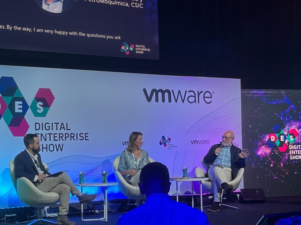 Rafael Mendías moderando sentado a la izquierda del mismo escenario, con micrófono y cuaderno, mientras intervienen los dos ponentes a su lado, ante paneles con los logotipos de DES y vmware