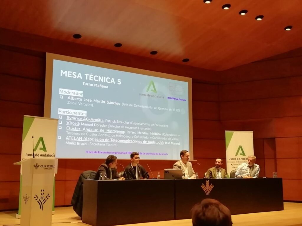 Auditorio con la pantalla mostrando la «Mesa técnica 5» del I Foro de Encuentro Empresarial FP/Empresa de la provincia de Granada, de RADFPDual Granada, con el Clúster Andaluz del Hidrógeno representado por Rafael Mendías Mellado, cofundador y tesorero y coordinador de VINLA, junto a Symrise AG-Armilla, Vircell y ATELAN; al fondo, cinco ponentes y roll-ups de la Junta de Andalucía y Caja Rural Granada